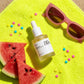 High-angle top-down flat lay photography of "Heartleaf 80% Soothing Ampoule" placed on a vibrant neon yellow beach towel. Surrounding the dropper bottle are fresh, juicy slices of red watermelon, star-shaped sprinkles, and retro pink sunglasses. Hard midday sunlight with crisp, dramatic shadows, fun and trendy Gen-Z aesthetic, 1:1 aspect ratio. (gemini-3.1-flash-image-preview)