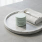 Angle : Vue à $30^\circ$ vers le bas.Scène : Le pot fermé est mis en scène sur un plateau rond en marbre blanc de Carrare aux veines grises très discrètes. À côté du produit, une serviette d'invité en coton nid d'abeille blanc est pliée proprement, surmontée d'un pinceau applicateur cosmétique à manche blanc.Fond & Lumière : Le plateau repose sur une surface en béton ciré gris clair. La lumière est douce, tamisée et diffuse, imitant la clarté d'un matin brumeux à travers une fenêtre de studio. (gemini-3.1-f