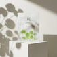 Angle : Centré, pile de face (Eye-level).  Scène : Le sachet de l'image image_cdfb9f.jpg se tient seul debout au centre d'un bloc cubique minimaliste laqué blanc mat. L'intérêt de l'image repose entièrement sur la mise en scène de la lumière.  Fond & Lumière : Fond monochrome blanc cassé. Une lumière dure traverse des feuillages botaniques de type Centella hors champ, projetant une ombre chinoise floue, poétique et parfaitement découpée de feuilles rondes sur le mur et en superposition sur le sachet blanc. 