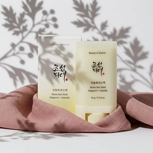 Une prise de vue frontale épurée et géométrique. Le stick solaire fermé et son packaging de l'image sont positionnés au centre Exact du cadre, debout. Leur base est délicatement enveloppée par un drapé de tissu de lin lourd couleur vieux rose poudré.  Détails : Fond monochrome blanc mat. Une lumière de studio directionnelle crue vient de biais de manière ciblée, projetant une ombre chinoise floue, poétique et parfaitement découpée de feuilles d'armoise et de baies sur le mur du fond et en superposition sur 