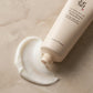 L'Angle : Gros plan macro de biais, à hauteur d'œil.  Le Fond : Le tube est posé à plat sur une surface en marbre mat beige. Juste à côté du bouchon, une magnifique ondulation (swatch) de crème blanche, onctueuse et veloutée est visible, montrant sa texture riche.  Le Prompt :  Macro commercial beauty shot of the Beauty of Joseon moisturizer tube lying flat on a smooth beige matte marble surface. Next to the opening cap, there is an elegant, thick, and velvety swirl swatch of the white face cream, showing i