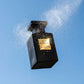 Style : Photographie en mouvement, atmosphérique.  Angle : Prise de vue en contre-plongée, avec un grand angle.  Fond : Un ciel d'été bleu clair et sans nuages.  Produit : Le petit flacon de parfum "Oud Kamare" noir opaque est comme suspendu dans les airs. Une légère brume d'eau est pulvérisée autour du bouchon, avec des particules d'eau fines qui attrapent la lumière, suggérant le moment où le parfum est pulvérisé.  Lumière : Lumière du soleil de midi, vive et directe, pour créer des reflets étincelants su