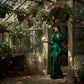 Un plan large d'un mannequin femme portant une robe couture structurée en soie verte émeraude profonde, tenant délicatement le flacon de image_3.png dans sa main gantée de satin vert. Elle se tient dans une orangerie ancienne, la lumière du soleil filtrant à travers le verre et les plantes de géranium suspendues. Son visage est partiellement ombragé. La plaque dorée de image_3.png capte une étincelle de soleil. L'ambiance est feutrée, mystérieuse. Style : Photographie de mode éditoriale, ambiance Renaissanc