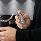 Un plan serré et intimiste centré sur les mains d'un mannequin homme vêtue d'un pull en maille de cachemire noir haut de gamme. Ses doigts, aux ongles soignés, tiennent délicatement le flacon de parfum transparent de image_19.png, extrait de la trousse de toilette visible en arrière-plan flou. La scène évoque le moment intime précédant la vaporisation du parfum. Style : Campagne publicitaire de beauté et de soin de la peau. (gemini-3.1-flash-image-preview)