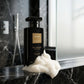 Un plan moyen de profil montrant uniquement l'imposant flacon de gel douche noir de image_221fc1.jpg posé sur le rebord en marbre noir d'une douche moderne. Une texture riche, vaporeuse et aérienne de mousse blanche onctueuse est délicatement déposée à sa base, contrastant avec l'opacité sombre de la bouteille. La lumière naturelle filtre à travers une vitre sablée. Style : Photographie lifestyle haut de gamme. (gemini-3.1-flash-image-preview)