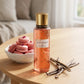 Un plan large du flacon de image_2288fe.jpg exposé sur une table en bois de santal clair et lisse, entouré d'une coupelle en céramique artisanale blanche contenant des macarons à la rose frais et des bâtons de vanille ouverts. Une lumière douce et naturelle traverse la scène, faisant vibrer l'éclat cuivré et rose du flacon du produit. Style : Photographie publicitaire de marque, naturelle et sensorielle. (gemini-3.1-flash-image-preview)
