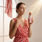 Un plan moyen d'un mannequin femme portant une robe d'été fluide rouge et blanche drapée, capturée de trois-quarts face dans un studio baigné d'une lumière blanche et ultra-douce. Elle tient le flacon de image_2ca76c.jpg avec grâce près de sa silhouette, les yeux clos dans une expression sereine de bien-être. Un rayon de soleil vient frapper doucement le bouchon en métal rouge de image_2ca76c.jpg. Style : Campagne de mode cinématographique. (gemini-3.1-flash-image-preview)