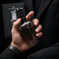 Un plan serré et intimiste centré sur les mains d'un mannequin homme au poignet orné d'une montre de luxe en acier brossé sombre. Ses doigts tiennent fermement et avec assurance le corps sculptural en verre fumé et métal noir du flacon de image_2de6bf.jpg, extrait de son coffret visible en arrière-plan flou. Le fond de l'image montre la texture noble d'une veste de costume sur mesure noire texturée. Style : Campagne publicitaire de mode et de parfum de luxe masculin. (gemini-3.1-flash-image-preview)