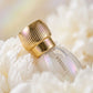 Un plan macro très rapproché concentré sur le bouchon cannelé en or poli et le col du flacon de image_2f4efb.jpg. Le produit est posé sur un lit de pétales frais de pivoine blanche texturée. Un éclairage irisé crée de fins reflets pastel nacrés sur le verre transparent et le métal doré de image_2f4efb.jpg. L'arrière-plan est un flou artistique (bokeh) très doux aux tons crème et or. Style : Photographie macro de beauté poétique. (gemini-3.1-flash-image-preview)