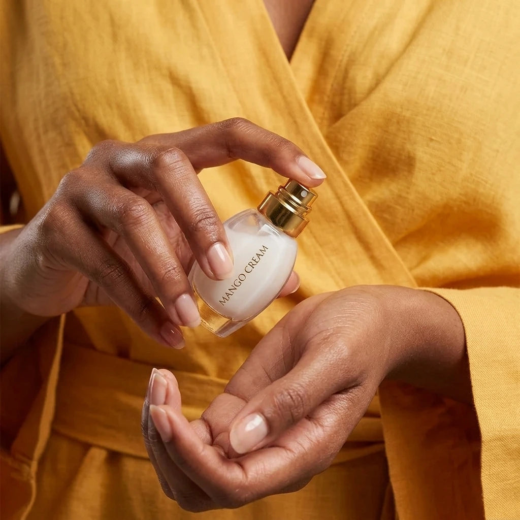 Un plan serré concentré sur les mains d'un mannequin femme à la peau satinée et hydratée. Ses doigts tiennent délicatement le flacon transparent de image_2f6169.jpg, s'apprêtant à appliquer une touche de ce musc crémeux au creux de son poignet. Le fond de l'image présente la texture floue d'un vêtement fluide en lin couleur jaune safran haut de gamme. Style : Campagne publicitaire de beauté et de soin intime. (gemini-3.1-flash-image-preview)