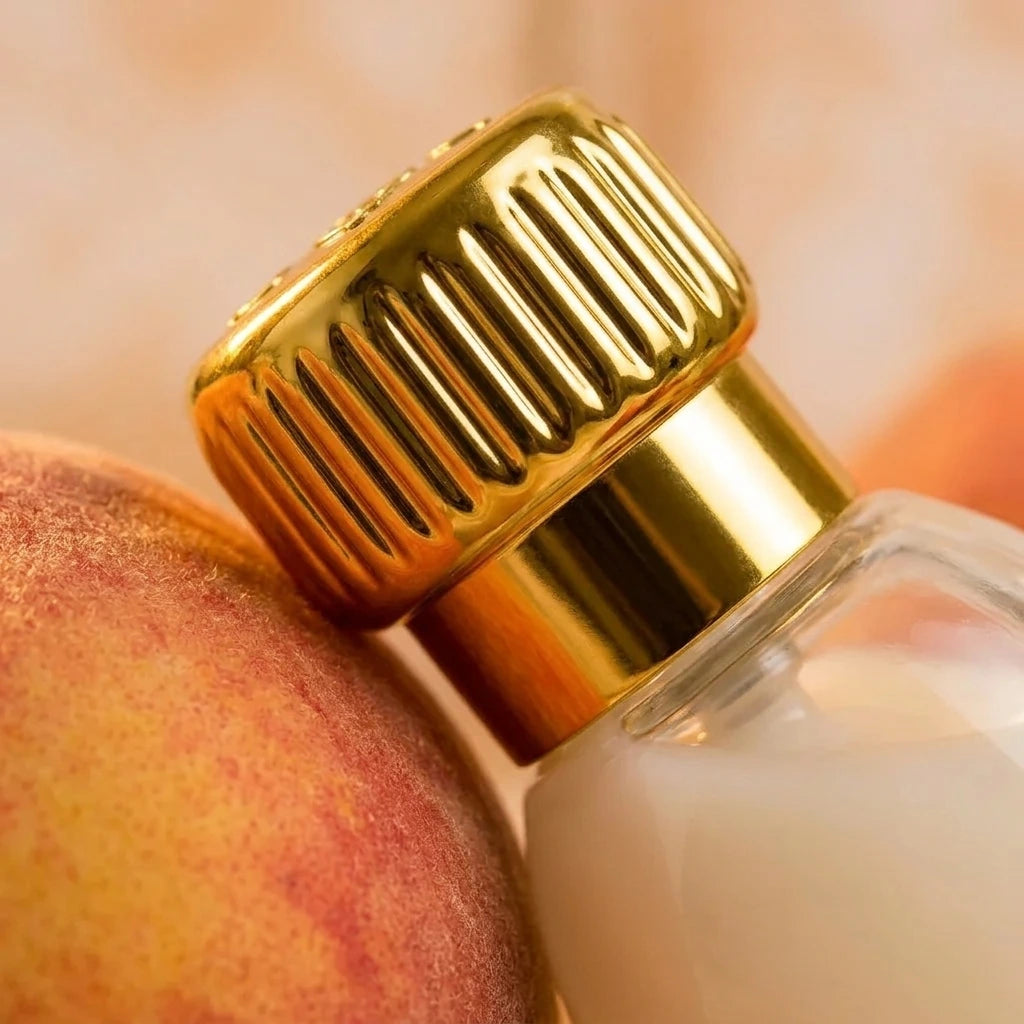 Un plan macro très rapproché concentré sur le bouchon en or poli strié et le col du flacon de image_3abf0c.jpg. Le produit est délicatement niché contre la peau veloutée d'une pêche de vigne fraîche et mûre. Un éclairage chaud crée de fins reflets dorés et ambrés sur les contours du verre transparent de image_3abf0c.jpg. L'arrière-plan présente un flou artistique (bokeh) très doux aux nuances crème. Style : Photographie de produit sensorielle. (gemini-3.1-flash-image-preview)