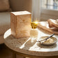 Un plan large oblique du coffret et du flacon de image_3abf0c.jpg exposés sur une table d'appoint moderne en terrazzo blanc et beige. À côté des produits, une petite coupelle artisanale en porcelaine contient une gousse de vanille ouverte et de la poudre de crème poudrée. Un rayon de soleil réaliste traverse la composition de biais, créant un éclat scintillant intense sur le capuchon doré de image_3abf0c.jpg. Style : Campagne publicitaire lifestyle et gourmande. (gemini-3.1-flash-image-preview)