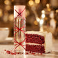 Angle : Gros plan macro, légèrement de profil.  Scène : Gros plan sur le flacon transparent de Pink Sugar Red Velvet de l'image image_7b5eae.jpg. Le ruban rouge croisé sur le verre capte la lumière. Juste à côté du flacon, une part de gâteau "Red Velvet" moelleuse et texturée est coupée, laissant voir sa couleur rouge profond et son glaçage blanc crème immaculé. Quelques miettes de gâteau rouges et des cristaux de sucre sont délicatement éparpillés à la base du parfum.  Fond : Arrière-plan flou (bokeh) de l