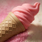 Angle : Gros plan macro serré, angle à $45^\circ$ braqué sur la jonction entre le cornet beige et la glace rose.Scène : Focus absolu sur les textures réalistes du flacon de "Cotton Blush" de l'image image_88f6ad.jpg. L'éclairage de studio (softbox très douce) enveloppe le capuchon rose mat pour en souligner les courbes parfaites sans créer de reflets durs. À côté de la base, quelques pincées de cristaux de sucre rose pailleté scintillent subtilement sous un spot directionnel.Fond : Arrière-plan flou de stud