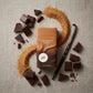 Angle : À plat ($90^\circ$ - Flat Lay de studio).Scène : Le flacon ondulé de l'image image_89601e.jpg est disposé horizontalement au centre. Autour de lui, une composition graphique impeccable : des morceaux de chocolat noir brisés, une gousse de vanille entière luisante et une fine traînée géométrique de sucre roux tamisé formant une courbe élégante.Fond : Une surface en tissu de lin texturé de couleur grège ou beige lin, éclairée par une lumière latérale douce. (gemini-3-pro-image-preview)
