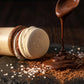 Angle : Gros plan macro très serré, incliné à 35 degrés, focus sur les macarons du capuchon.  Scène : Le flacon de l'image image_8bb442.jpg est posé en plein cœur d'un plan de travail de pâtisserie en ardoise sombre. Juste à côté, un filet de chocolat noir chaud et brillant est coulé à la cuillère, figeant ses vagues satinées. Quelques cristaux de fleur de sel et de la poudre de cacao brute sont saupoudrés à la base, rappelant le fini mat du flacon.  Fond & Éclairage : Arrière-plan sombre et texturé (bois d