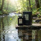 Concept de shooting : Intégrer un mouvement naturel (l'eau ou le vent) pour casser la monotonie d'un shooting classique.  Le Prompt à copier :  Une photographie e-commerce créative et vivante. Le flacon de parfum "BLACK EMPIRE" et sa boîte noire sont installés sur une avancée de pierre plate au bord d'un ruisseau forestier calme. L'eau limpide coule et ondule doucement au premier plan, créant des cercles concentriques qui reflètent la silhouette des produits. Une branche fine avec quelques feuilles vertes f
