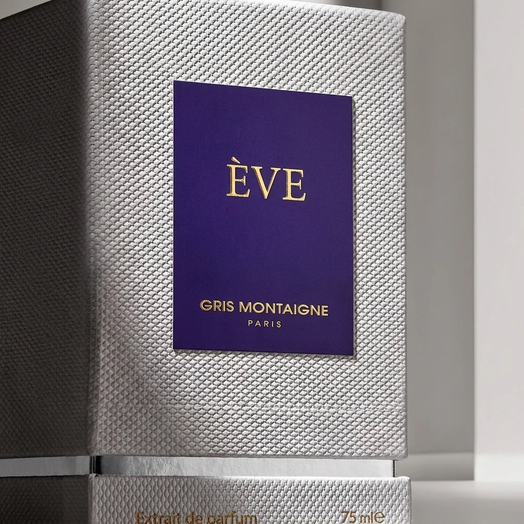 Un plan macro très serré et frontal sur la boîte texturée de image_3.png, en studio. L'éclairage est rasant pour accentuer la trame gaufrée du coffret. Le centre de l'image est la plaque violette avec le texte 'ÈVE GRIS MONTAIGNE PARIS' doré, qui est parfaitement net. La base argentée et l'inscription 'Extrait de parfum' sont visibles. Style : Photographie d'architecture d'intérieur minimaliste. (gemini-3-pro-image-preview)