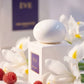 Un plan serré de profil du flacon blanc épuré de image_3.png. Le capuchon sphérique blanc est le point central de la netteté. Il est entouré de véritables fleurs d'iris blancs et de quelques baies de framboise. La lumière est diffuse et douce, presque éthérée. La plaque violette est visible en arrière-plan flou (bokeh). Style : Photographie de beauté conceptuelle. (gemini-3-pro-image-preview)
