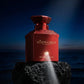Un plan large en contre-plongée (vue basse) plaçant le flacon de image_27.png sur un piédestal de roche volcanique noire poreuse. Le fond est un dégradé de bleu nuit et de noir évoquant un crépuscule sur la mer Méditerranée. Un faisceau de lumière blanche et directionnelle frappe directement le logo doré au bas de image_27.png, le rendant parfaitement net, lisible et haut de gamme. Style : Photographie de produit dramatique et conceptuelle. (gemini-3-pro-image-preview)