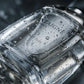 Un plan macro très serré concentré sur le verre lourd du flacon de parfum transparent de image_19.png. De micro-gouttelettes d'eau pure perlent de façon réaliste sur la paroi lisse du verre, faisant écho à la fraîcheur de la mousse de douche. L'arrière-plan présente un flou artistique (bokeh) très sombre aux nuances grises et argentées. Style : Photographie de produit sensorielle et tactile. (gemini-3-pro-image-preview)