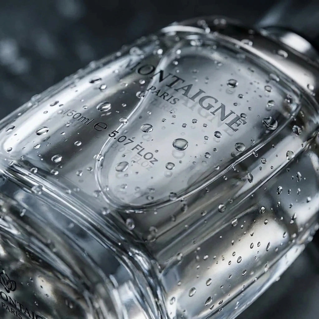 Un plan macro très serré concentré sur le verre lourd du flacon de parfum transparent de image_19.png. De micro-gouttelettes d'eau pure perlent de façon réaliste sur la paroi lisse du verre, faisant écho à la fraîcheur de la mousse de douche. L'arrière-plan présente un flou artistique (bokeh) très sombre aux nuances grises et argentées. Style : Photographie de produit sensorielle et tactile. (gemini-3-pro-image-preview)