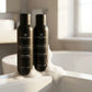Un plan moyen de profil montrant uniquement les deux flacons cylindriques noirs mats et satinés de image_19.png posés sur le rebord d'une baignoire en céramique moderne. Une texture riche, vaporeuse et aérienne de mousse de douche blanche onctueuse est délicatement déposée à leur base. La lumière naturelle du matin inonde la scène. Style : Photographie lifestyle haut de gamme. (gemini-3-pro-image-preview)