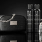 Un plan moyen de face respectant la composition complète de image_19.png : la trousse noire embossée et sa chaîne argentée sont installées à gauche, tandis que les deux flacons de soin noirs mats et le flacon de parfum transparent sont alignés avec élégance à droite sur une surface laquée noire et lisse. Le sol crée un reflet miroir net et impeccable sous chaque élément. Style : Packshot commercial haut de gamme. (gemini-3-pro-image-preview)