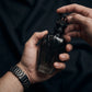 Un plan serré et intimiste centré sur les mains d'un mannequin homme au poignet orné d'une montre de luxe en titane brossé noir. Ses doigts géométriquement positionnés tiennent fermement et avec assurance le corps sculptural en verre fumé et métal noir du flacon de image_2ee9ff.jpg, s'apprêtant à soulever le capuchon en couronne noire. Le fond est un tissu technique flou noir anthracite. Style : Campagne publicitaire éditoriale de mode. (gemini-3-pro-image-preview)
