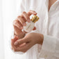 Un plan serré concentré sur les mains d'un mannequin femme à la peau douce et naturelle, portant une chemise fine en lin blanc. Ses doigts aux ongles impeccables tiennent délicatement le flacon ovoïde de image_2f4efb.jpg, s'apprêtant à appliquer une touche de ce musc crémeux sur son poignet. L'arrière-plan est baigné d'une lumière blanche et floue ultra-douce. Style : Campagne publicitaire de soin et de beauté intime. (gemini-3-pro-image-preview)