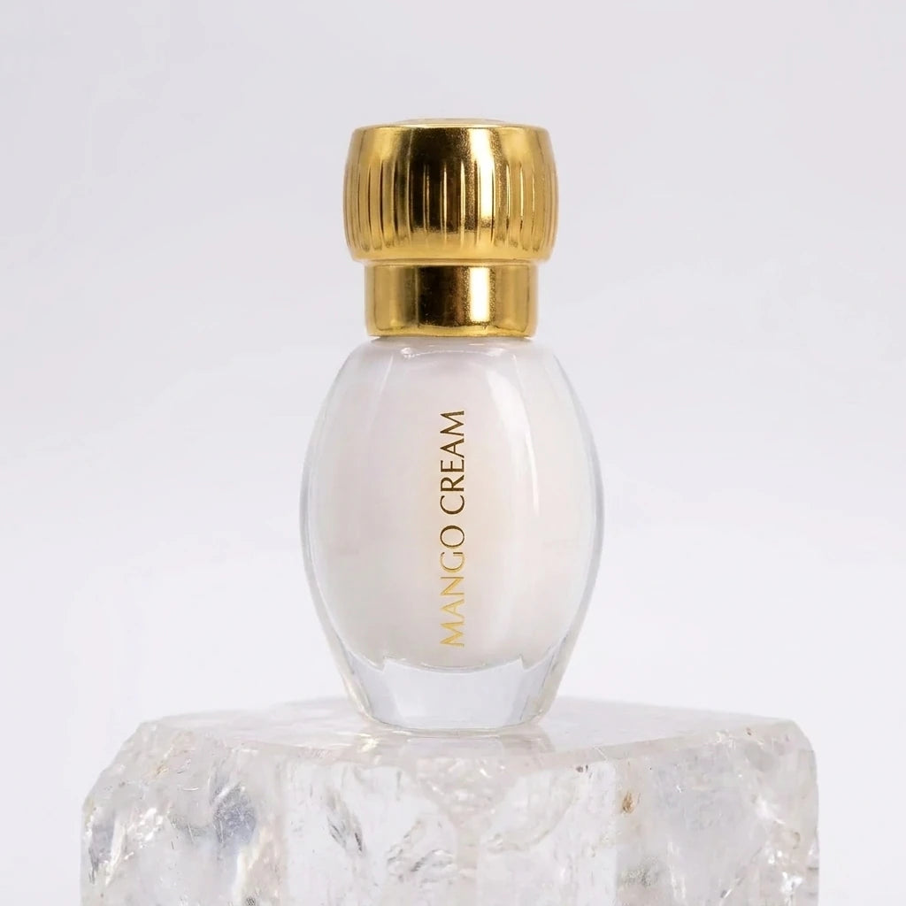 Un plan moyen frontal du petit flacon ovoïde de image_2f6169.jpg isolé, posé sur un bloc de quartz cristallin transparent. Une lumière douce traverse la scène par l'avant, révélant la texture blanche et laiteuse du jus à travers le verre transparent et faisant briller l'inscription verticale jaune-dorée 'MANGO CREAM' de image_2f6169.jpg. Le bouchon doré cannelé capte un éclat lumineux linéaire parfait. Style : Photographie de produit haut de gamme et épurée. (gemini-3-pro-image-preview)