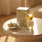 Un plan large oblique du coffret et du flacon de image_2fbbbb.jpg disposés sur une table d'appoint moderne en terrazzo beige. À côté des produits, une petite coupelle en porcelaine blanche contient des éclats de pistaches grillées et une gousse de vanille entrouverte. Un rayon de soleil réaliste de l'après-midi traverse la scène, créant un éclat brillant sur le capuchon doré de image_2fbbbb.jpg. Style : Campagne publicitaire lifestyle et sensorielle. (gemini-3-pro-image-preview)
