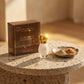 Un plan large oblique du coffret et du flacon de image_2fc3fc.jpg disposés sur une table d'appoint moderne en terrazzo beige et marron. À côté des produits, une petite coupelle artisanale en céramique contient de la poudre de fève tonka fine et des éclats de sucre roux. Un rayon de soleil réaliste de fin de journée traverse la scène, créant un éclat brillant sur le capuchon doré de image_2fc3fc.jpg. Style : Campagne publicitaire lifestyle et sensorielle. (gemini-3-pro-image-preview)