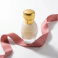 Un plan moyen en plongée à 60 degrés du flacon de image_30acb9.jpg délicatement posé sur une surface laquée blanche impeccable. Un ruban de soie fluide couleur vieux rose ondule de manière asymétrique autour de la bouteille. La lumière rasante souligne la rondeur parfaite du flacon de image_30acb9.jpg et l'éclat miroir de son bouchon métallique doré. Style : Éditorial de beauté épuré. (gemini-3-pro-image-preview)