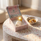 Un plan large oblique du coffret ouvert et du flacon de image_3aa887.jpg exposés sur une console moderne en terrazzo blanc et rose pastel. À côté des produits, une petite coupelle artisanale en porcelaine contient une gousse de vanille entrouverte et de la poudre de sucre roux. Un rayon de soleil réaliste traverse la scène de biais, créant un éclat scintillant intense sur la couronne dorée de image_3aa887.jpg. Style : Campagne publicitaire lifestyle. (gemini-3-pro-image-preview)