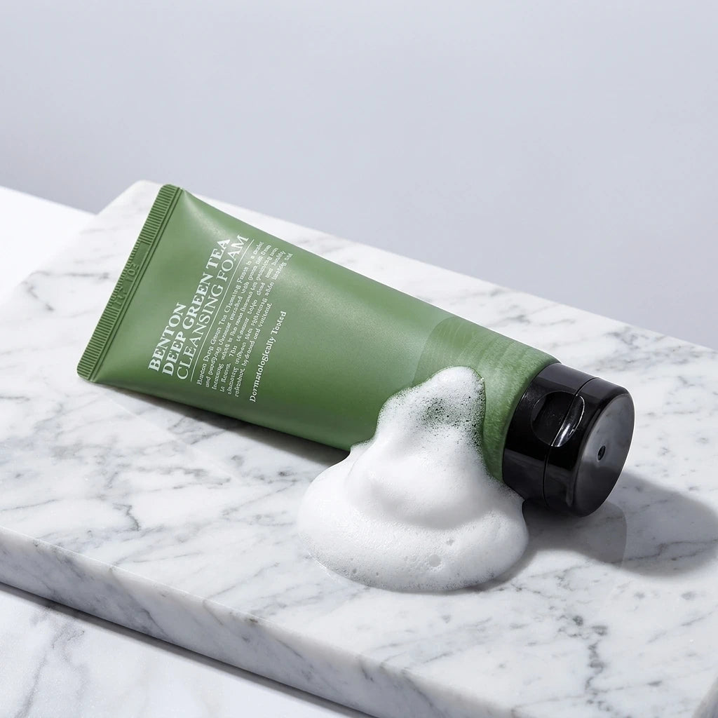 Macro-photographie publicitaire de produit cosmétique. Le tube vert de "Benton Deep Green Tea Cleansing Foam" est allongé sur une surface de marbre blanc immaculé et veiné. À côté du bouchon, une généreuse noisette de mousse nettoyante blanche, incroyablement dense, micro-bullée et aérienne, est artistiquement étalée. Éclairage de studio professionnel, propre, contrastes doux, ombres subtiles. Look ultra-propre, luxueux et minimaliste. (gemini-3-pro-image-preview)
