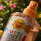 An extreme macro close-up photograph of the Chupa Chups Tutti Frutti Brume Parfumée (Body Mist) bottle, focusing intensely on intricate textures. The camera is super close, showing the palpable, slightly grainy paper texture of the 'TUTTI FRUTTI' label and the fine ink dot patterns of the colorful (pink, orange, blue, green) letters. The transparent plastic of the bottle surface reveals minute scratches, imperfections, microscopic dust, and small air bubbles suspended in the shimmering pink-orange liquid in