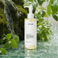 La Cascade Douce (Aqueux & Naturel)Angle : Trois Quarts DynamiquePrompt : A refreshing commercial product shot of the "Heartleaf Pore Control Cleansing Oil" dropper bottle. The bottle stands on a polished, natural green marble surface. A gentle, controlled stream of water cascades down a raw moss-covered rock formation behind it, pooling slightly around the base without submerging it. Fresh heartleaf greenery and tiny white flowers cluster near the moss. The lighting is bright and diffused, highlighting the