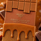 Une photo macro carrée à 1200x1200px. Un gros plan extrême sur l'étiquette centrale de la bouteille d'Arabiyat Sugar 'TOFFEE GANACHE' d'image_1.png. La texture de la tablette de chocolat au caramel au-dessus et la base granuleuse en-dessous sont nettes. La mise au point est si précise sur les mots 'ARABIYAT' et 'TOFFEE GANACHE' qu'on peut en voir les détails. L'arrière-plan est un flou bokeh de caramel liquide chaud et de morceaux de chocolat fondu, suggérant une cuisine de pâtissier. La lumière est douce e