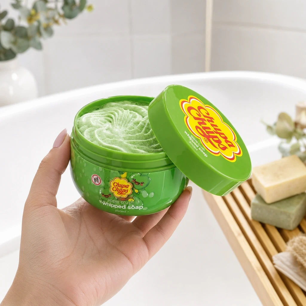 A commercial frontal photograph (angle frontal) in a bright bathroom. A woman's hand, with manicured nails, holds the 200ml Chupa Chups - Apple Crush - Whipped Soap pot. The yellow lid is partially unscrewed and tilted, revealing the fluffy, green-swirled whipped soap inside. The background shows a white bathtub with some unbranded artisan soaps on a wooden rack. Light reflects on the product label. (gpt-image-2)